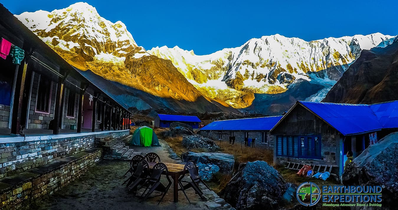 Blue-roofed stone lodges and a dining area beneath snow peaks lit by the morning sun
